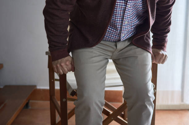 A mature man has difficulty getting up from an armchair in the living room of his home. Horizontal photo. Selective blur. A mature man has difficulty getting up from an armchair in the living room of his home. standing had to knee pose stock pictures, royalty-free photos & images