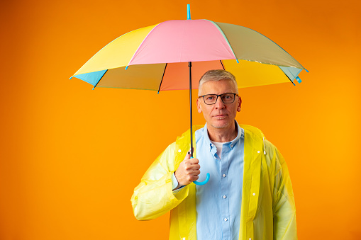 Senior man wearing yellow plastic overcoat and holding rainbow umbrella over yellow background, close up