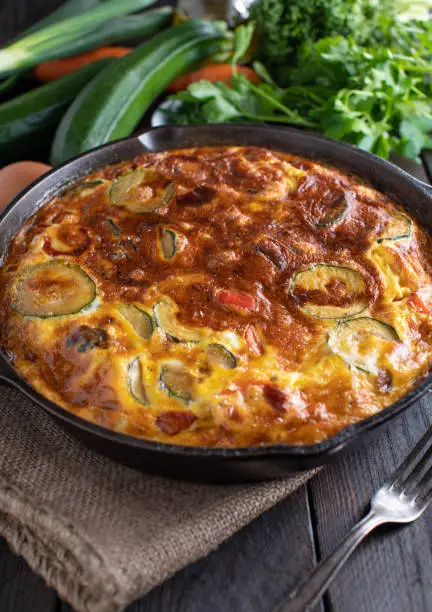 Homemade fresh cooked healthy low carb fritatta with zucchini, tomatoes, bell peppers and red onions. Served in a rustic cast iron pan on wooden table. Ready to eat.