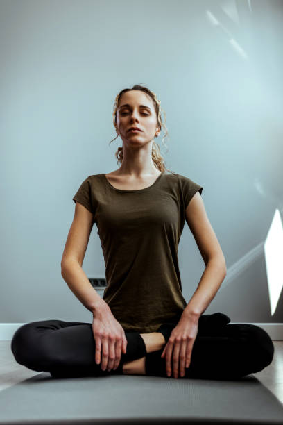 Young woman sitting in lotus position on exercise mat with her eyes closed and meditating in the living room Happy young woman practicing her yoga routine at home yoga studio photos stock pictures, royalty-free photos & images