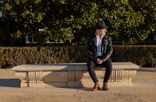 Adult man in hat and leather jacket sitting on bench in park during a sunny day. Madrid, Spain