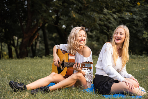 Portrait of caucasian young women sitting in the park outdoor and playing a guitar sing a song together with happiness