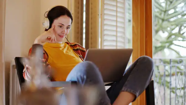 Stock image of a dark-haired young woman reclining at home with her laptop & headphones,  she’s working casually.
