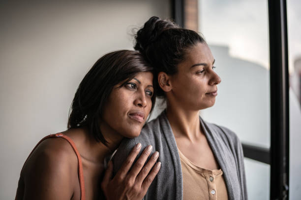 Contemplative lesbian couple looking through window at home Contemplative lesbian couple at home sad gay stock pictures, royalty-free photos & images