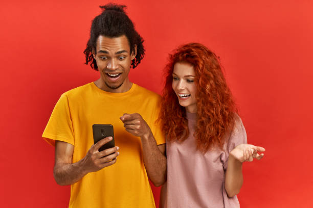istock Optimistic young ginger female and Afroman users of smart technology, feel good from successful cellphone updating, gaze at screen, check article, wear casual clothes, stand over red background 1385947034