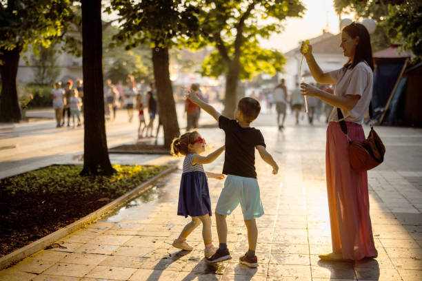 Mother standing in park and blowing bubbles with bubble wand while two kids catching them