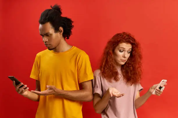 Connection error. Black millennial man and redhead woman looking at smartphones, annoyed with discharged or broken cellphones, express surprisement and annoyance, received bad news in message