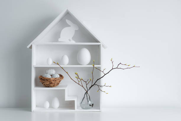 Home minimalist interior with Easter decor. Branches with budding buds in a glass vase on shelves in the shape of a house,
