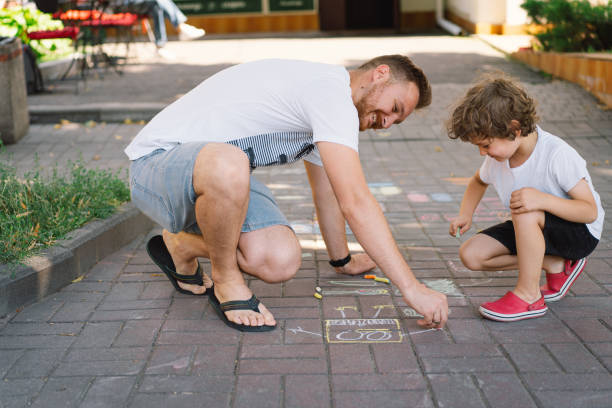 маленький дошкольник с отцом рисует разноцветными мелами на земле. - child art paint humor стоковые фото и изображения