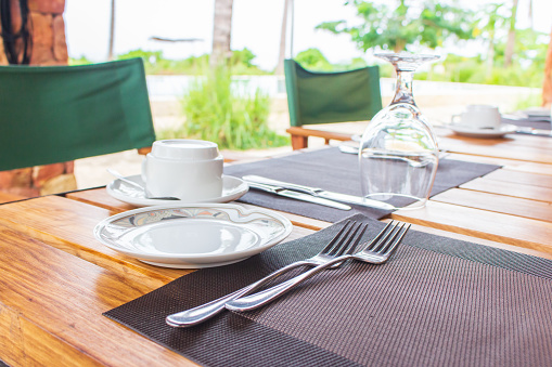 Table setting in tropical resort. Table set near resort pool. Cutlery on table of outdoor restaurant. Wine glass and cup on dinner table in luxury villa. Restaurant near swimmimg pool with palm trees.