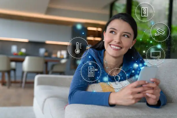Portrait of a Latin American woman controlling features of her smart home from her cell phone while lying on the couch and smiling - technology concepts