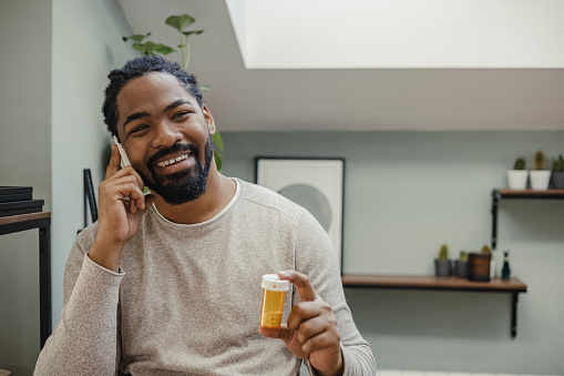 African American man is at home. He is communicating with the doctor via mobile phone while holding a bottle of medicine in his hand.
