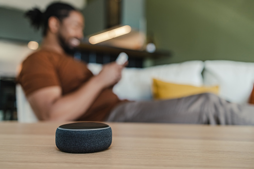 Young African American man sitting on the couch and looking at his smart phone. Smart speaker is placed on a table in the living room.