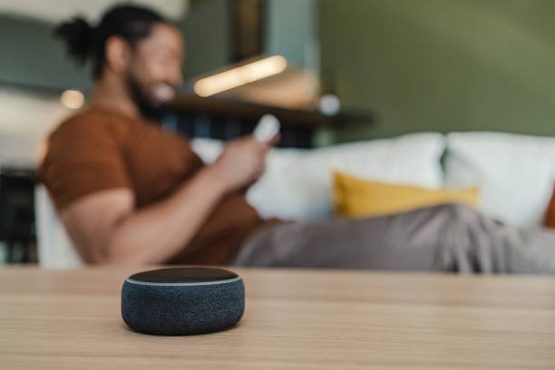 Smart speaker on the table in the living room. Young African American man sitting on the couch and looking at his smart phone. Smart speaker is placed on a table in the living room. virtual assistant stock pictures, royalty-free photos & images