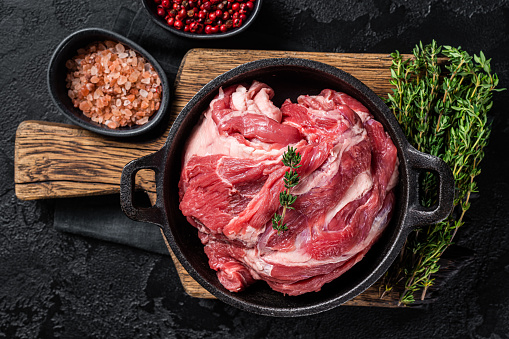 Boneless lamb neck, raw meat in a skillet with herbs. Black background. Top view.