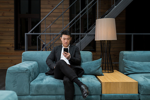 Successful Asian businessman manager, in a black business suit, uses the phone for video calls and conferences relaxing in a restaurant or hotel, and in a stylish office explains