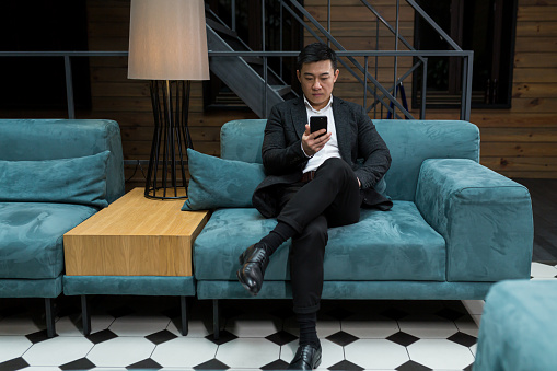 Successful Asian businessman manager, in a black business suit, uses the phone for video calls and conferences relaxing in a restaurant or hotel, and in a stylish office explains