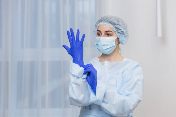 Young female doctor prepares for surgery, wears blue surgical gloves, in a white coat and mask, looks at the camera Young female doctor prepares for surgery, wears blue surgical gloves, in a white coat and mask, looks at the camera protective workwear stock pictures, royalty-free photos & images
