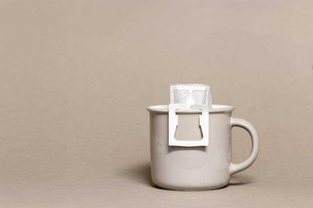 istock Drip coffee bag with ground coffee in big cup, package with paper bag drip coffee filter for brewing on beige color background, side view with copy space, monochrome color 1386010075