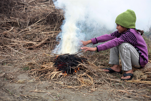 Elementary age poor girl of Indian ethnicity sitting outdoor in nature and warming hands using fire.