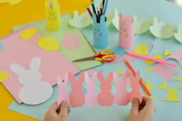 Photo of a close-up of a schoolboy who holds a garland of pink bunnies in his hands. Easter crafts, craft tools and materials on a table. Festive spring crafts concept