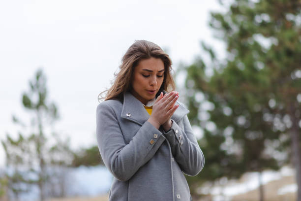 A young beautiful woman is blowing into her hands because it is cold outside. She is wearing a grey coat and is in a forest.