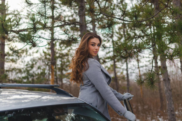 junge schöne frau in einem grauen mantel, die auf ihrem auto in einer nicht-städtischen umgebung posiert - winter non urban scene snow tree stock-fotos und bilder