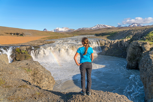 Rear view of young women standing at scanic landscape at Gýgjarfoss waterfall, river flowing from melting snow at the Kerlingarfjöll mountain range against clear blue sky.