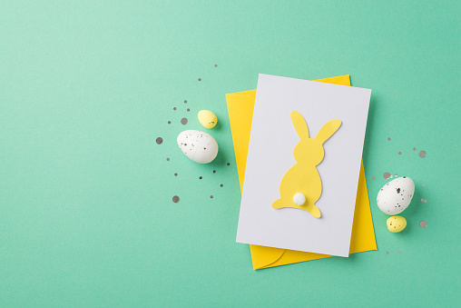 Top view photo of easter decorations shiny confetti yellow envelope paper card with easter bunny silhouette and easter eggs on isolated teal background with copyspace
