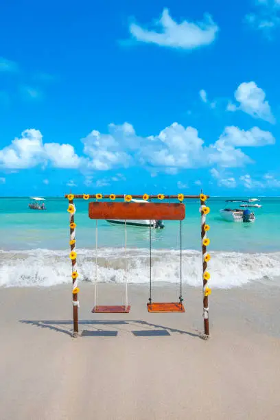Two seat swing decorated with sunflower flowers on a beautiful beach of fine sand and cyan-toned sea on a beautiful blue sky day. Landscape of Maragogi beach, Alagoas state, Brazil.