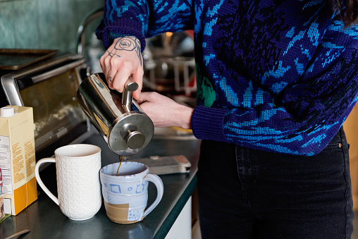 Young woman enjoying simple pleasures at home, making coffee. She is in her late twenties, wearing casual clothes. No face. Apartment is very lived in and cosy. Horizontal waist up indoors shot.