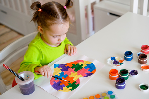 Beautiful drawing with a heart made of puzzle pieces as a sign of support for people suffering from autism. Baby paints a sheet of paper with watercolor. Sweet Girl with ponytails sitting at the table