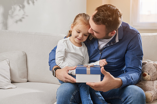Smiling little girl giving present in box for dad while sitting on sofa and celebrating Fathers Day at home