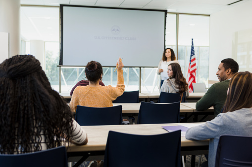 A rear view of a classroom full of diverse students taking an American citizenship class.  One of the students, a woman, raises her hand to ask a question.
