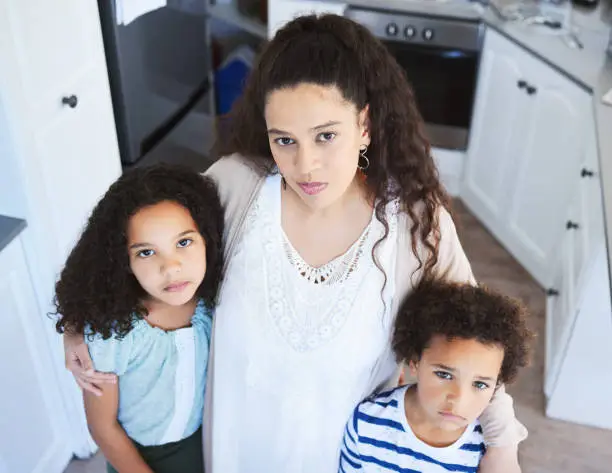 Photo of Shot of a young mother holding her children while standing in the kitchen at home