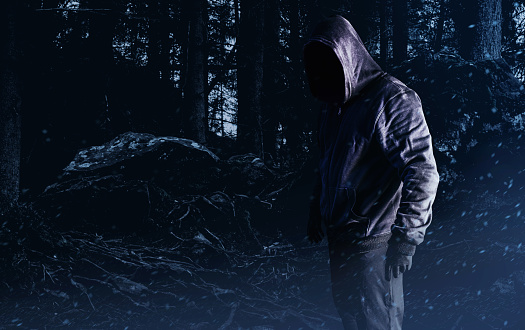 Photo of scary horror stranger stalker man in black hood and clothing on forest background.