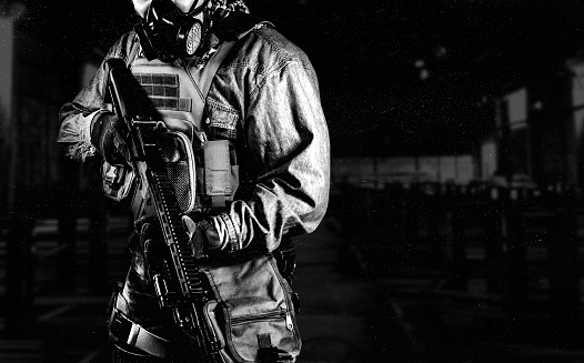 Black and white photo of urban soldier in tactical military outfit and gas mask standing with rifle and gas mask on dark factory background.