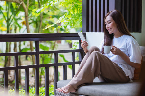 istock Portrait image of a beautiful young asian woman holding and using mobile phone while drinking coffee and sitting on sofa at home 1386105325