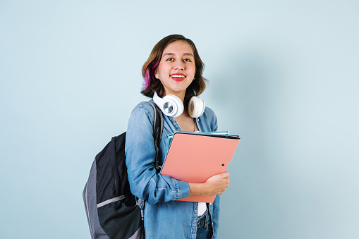 Young Hispanic student woman listen music with headphones and holding computer over isolated blue background
