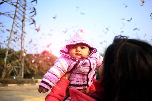 Young woman playing with her baby daughter against birds background outdoors in the park.