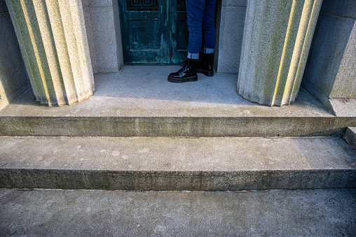 Ancient stone pillars on either side of the door to a mausoleum with one lone mourner at the door. Stone steps and detailed elegant architecture, mourner wears black shoes, shot in natural light with copy space and one person, feet only.