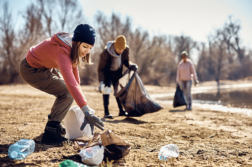 Teenage girl picking up garbage while cleaning the beach with group of volunteers.