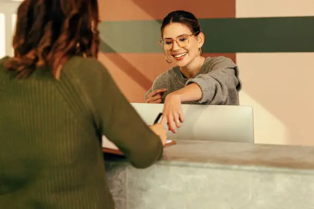 Easygoing receptionist assisting a woman with signing in to an office. Friendly receptionist showing a woman where to sign on a clipboard. Young woman working at the front desk of a convention centre.