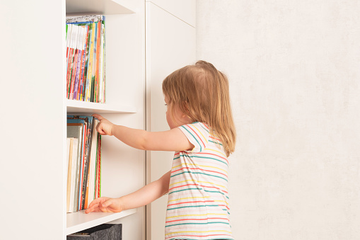 Little girl chooses a book in a bookcase at home. The child looks through the books in the library, deciding which one to take to study. Children's creativity and education