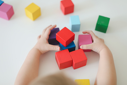 Top view on child hands playing and building with colorful wooden toy bricks on white wooden table. Early learning and development.