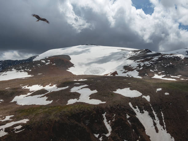 istock Bright atmospheric scenery on top of spotted snow mountain ridge under dark clouds. Beautiful landscape with fly eagle above mountain range with sunlight under stormy dense clouds. 1386192011