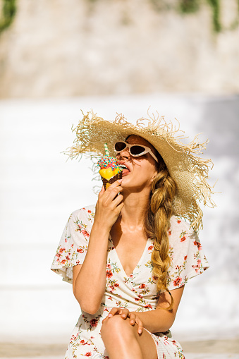 Gorgeous young woman sitting outdoors on stairs enjoying while eating ice cream at sunny day. She is tourist on vacation on Greek islands and looks happy and carefree