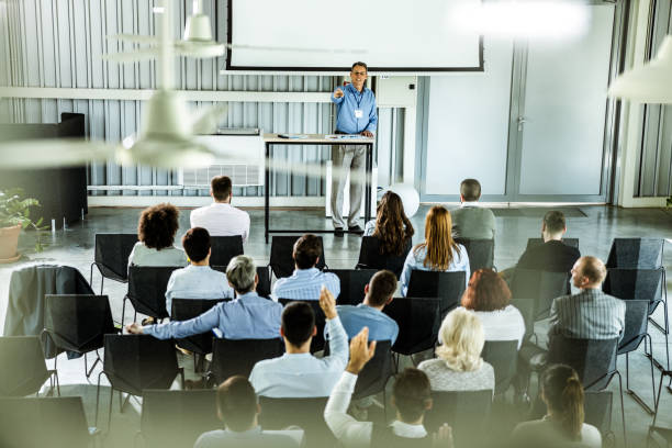 Large group of business people listening to public speaker on a seminar in board room while some of them are raising their hands to ask a question.
