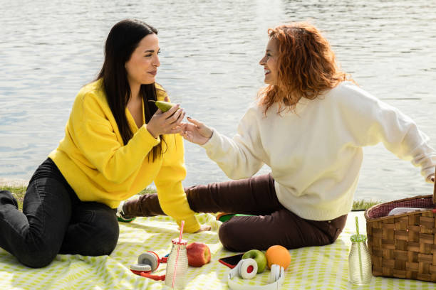 湖畔で春の新鮮なフルーツピクニックを共にし、日差しの中でおしゃべりをしながら微笑む女友達たち。 - two pears ストックフォトと画像
