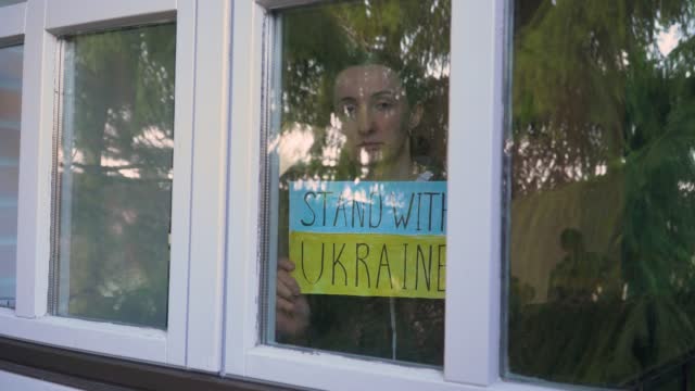 Scared woman showing through window placard with inscription Stand with Ukraine on yellow blue flag background